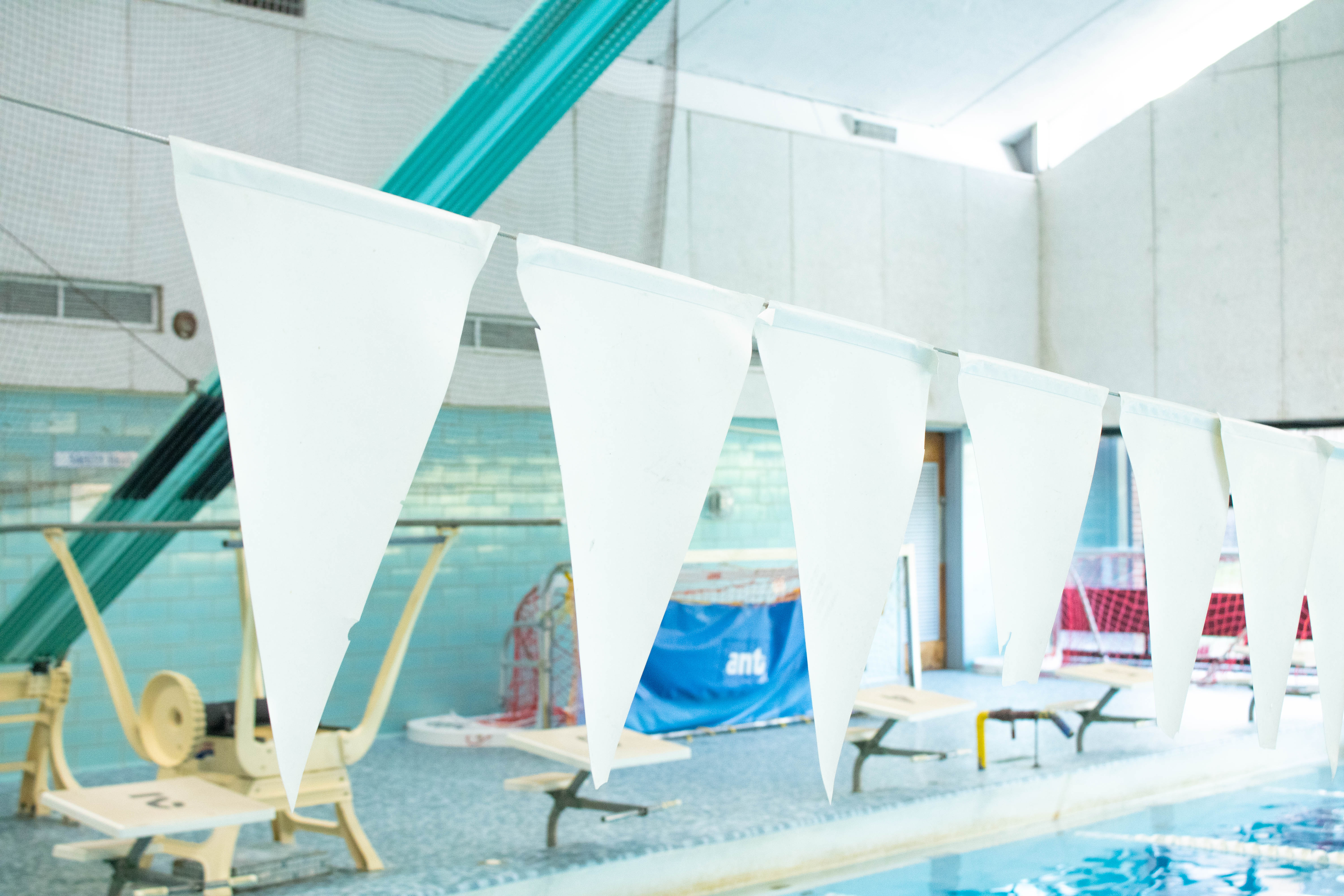 Image of white flags with a teal diving board in the background with white blocks below it and water polo equipment behind it.